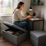 Woman working at a small home office desk while sitting on a storage ottoman with the lid open showing organized office supplies inside