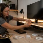 Woman installs LED strip lights behind a monitor and under a desk shelf to create ambient lighting in a small home office.