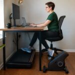 Office worker typing at a desk in a small home office while using an under desk treadmill and a bike pedal exerciser.